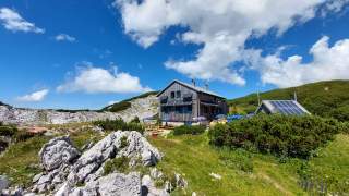 Berghütte mit Solarpaneelen und Außensitzbereich in einer alpinen Landschaft unter blauem Himmel mit weißen Wolken.