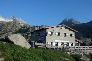Steinerne Berghütte mit blauen Fensterläden vor einer Berglandschaft unter klarem, blauem Himmel.