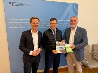 Drei Männer stehen lächelnd vor einer blauen Wand mit dem Logo des Bundesministeriums für Umwelt; zwei von ihnen halten Broschüren in den Händen.