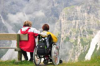 Eine Rollstuhlfahrerin und eine Frau auf einer Bank schauen von einem Aussichtspunkt auf eine imposante Felswand auf der anderen Talseite.
