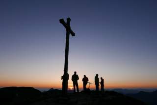 Scherenschnittartig vier Wanderer und ein großes Gipfelkreuz bei Sonnenaufgang.