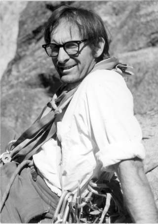 Schwarz-weiß-Foto eines Mannes mit Brille und Kletterausrüstung, der lächelnd vor einem Felsvorsprung sitzt.