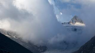 Berggipfel mit Schnee und Gletscher, teils von dichten Wolken umgeben und von blauem Himmel im Hintergrund.