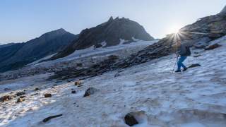 Ein Wanderer mit Trekkingstöcken geht bei Sonnenaufgang über einen schneebedeckten Hang in den Bergen, im Hintergrund sind spitze Berggipfel zu sehen.