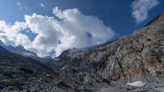 Berglandschaft mit steinigem Untergrund und kahlen Felswänden unter einem blauem Himmel mit großen weißen Wolken.