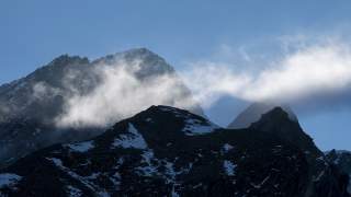 Dunkle, schneebedeckte Berggipfel mit aufsteigendem Nebel vor klarem blauem Himmel.