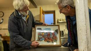 Zwei ältere Männer betrachten lächelnd ein gerahmtes Foto einer großen Gruppe von Menschen in Sportkleidung. Sie stehen in einem rustikalen Raum mit Küchenutensilien im Hintergrund.