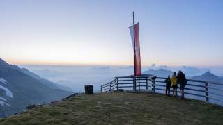 Drei Personen schauen bei Sonnenaufgang von einem Berggipfel mit einer Österreich-Flagge auf eine neblige Berglandschaft. Ein Holzzaun umgibt den Aussichtspunkt.