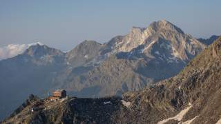 Berglandschaft bei klarem Himmel mit einem Berghaus auf einem felsigen Gipfel im Vordergrund und hohen, teilweise schneebedeckten Bergen im Hintergrund.