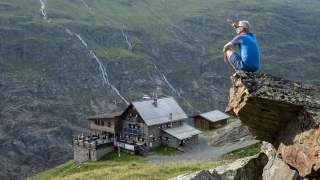Ein Mann in blauer Sportkleidung sitzt auf einem Felsen und schaut auf eine Berghütte in einem grünen, felsigen Tal mit kleinen Wasserfällen im Hintergrund.