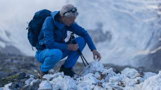 Ein Wanderer in blauer Outdoor-Kleidung kniet auf felsigem Gelände und stapelt Steine zu einem kleinen Haufen, mit verschneiten Bergen im Hintergrund.