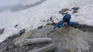 Ein Bergsteiger mit Rucksack und Sonnenbrille klettert an einem felsigen, schneebedeckten Hang und greift nach dem Ende einer Holzleiter. Nebel und Geröll sind im Hintergrund sichtbar.