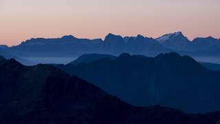 Silhouetten von Bergketten vor einem pastellfarbenen Himmel bei Dämmerung, mit leichtem Nebel in den Tälern.