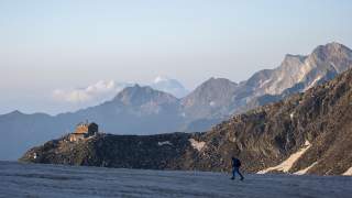Ein Wanderer läuft über eine Schneefläche in den Bergen, im Hintergrund ist eine Berghütte auf einem Felsen und eine Bergkette unter klarem Himmel zu sehen.
