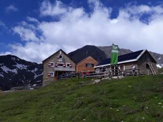 Berghütte mit mehreren Gebäuden, umgeben von grüner Wiese und Bergen mit Schneeflecken unter blauem Himmel mit Wolken.