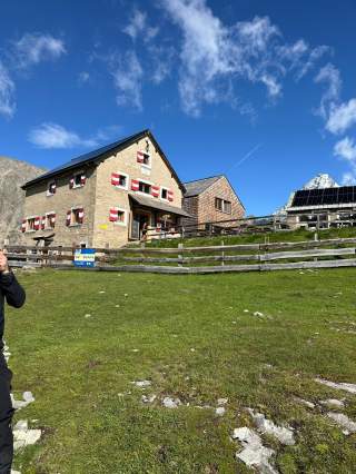 Altes Alpenhaus mit rot-weißen Fensterläden auf einer Grünfläche, dahinter ein weiteres Gebäude mit Solarpanelen und schneebedeckter Berggipfel unter blauem Himmel.