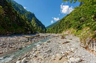 Ein schmaler Flusslauf in einem Tal, die Hänge sind bewaldet & grün. Blauer Himmel mit einigen Wolken.