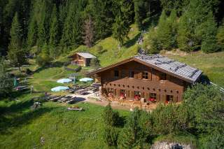 Hütte mit Blumen an den Fenstern umgeben von grünen Wiesen, Tischen mit Sonnenschirmen und dichtem Nadelwald im Hintergrund.