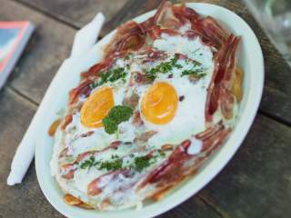 Ein Teller mit zwei Spiegeleiern, umgeben von gebratenem Speck und garniert mit frischen Kräutern, steht auf einem Holztisch.
