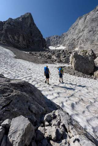 Zwei Personen queren Schneefeld in felsigem Berggelände