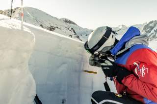 Person in winter gear and helmet examining a snow profile in a dug-out snow pit, surrounded by snowy mountains.