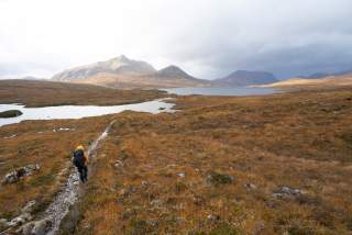 Eine Person mit gelber Jacke wandert auf einem schmalen Pfad durch eine herbstliche Moorlandschaft mit Bergen und Seen im Hintergrund.