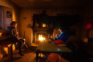 Zwei Personen sitzen in einer einfachen, schwach beleuchteten Hütte am Kaminfeuer; Kleidung hängt über dem Feuer zum Trocknen, eine Person trägt eine Stirnlampe.
