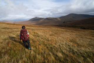 Person mit großem Rucksack wandert durch eine weite, grasbewachsene Hügellandschaft, im Hintergrund Berge und bewölkter Himmel.
