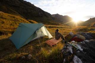 Zelt und Campingausrüstung auf einer Wiese in den Bergen bei Sonnenaufgang, eine Person sitzt auf einer Isomatte und blickt ins Tal.