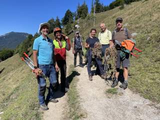 Sechs Menschen stehen auf einem Bergweg, halten Werkzeuge und Seile, tragen Outdoor-Kleidung und lächeln in die Kamera; im Hintergrund Berge und Bäume.