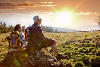 Ein älteres Paar macht beim Wandern eine Pause auf einem Stein. Sie schauen sitzend in die Abendsonne.