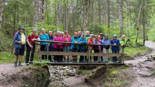 Eine Gruppe älterer Menschen steht mit Wanderkleidung und Rucksäcken auf einer kleinen Holzbrücke im Wald.