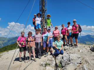 Gruppe von Wanderern steht bei sonnigem Wetter an einem Gipfelkreuz auf einem Berg, im Hintergrund Berge und blauer Himmel mit wenigen Wolken.