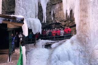 Gruppe von Menschen steht auf einem Steg in einer Eishöhle, umgeben von großen Eiszapfen und gefrorenen Wasserfällen.