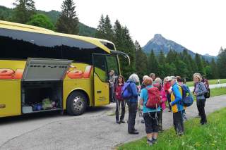 Eine Gruppe älterer Wanderer mit Rucksäcken und Wanderstöcken steht vor einem gelben Reisebus, im Hintergrund Berge und Wald.