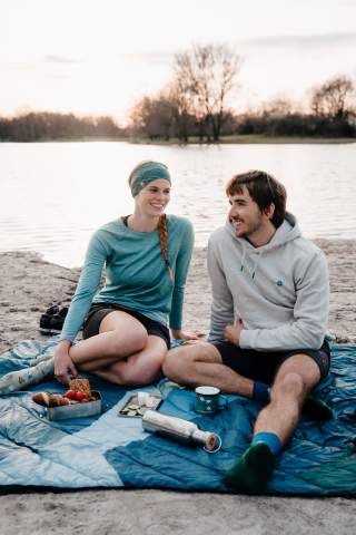 Dame und Herr sitzen lachend an einem See. Beide tragen schöne Artikel der neuen DAV-Kollektion.