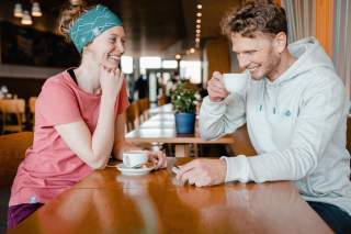 Dame und Herr sitzen sich lachend in einem Cafe gegenüber. Beide sind mit Artikeln der neuen DAV-Kollektion eingekleidet.