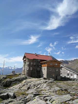 Steinernes Berghütte mit rotem Dach auf felsigem Untergrund, im Hintergrund Berge und blauer Himmel mit Wolken.
