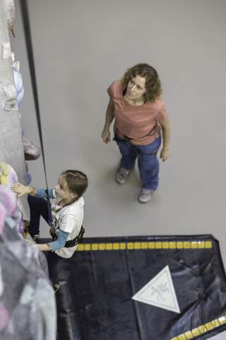 Ein Kind klettert an einer Kletterwand, während eine Frau am Boden stehend zuschaut und sichert.