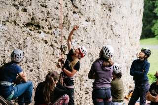 Mehrere Menschen mit Kletterausrüstung und Helmen stehen und sitzen vor einer Felswand, eine Person zeigt nach oben und erklärt etwas.