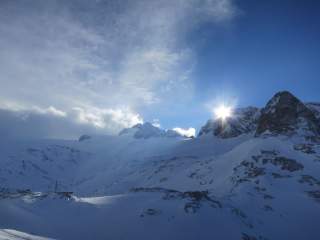 Tiefverschneite, sehr schattige und einsame Hochgebirgslandschaft im Hochwinter. Hinter einem Berg blitzt die Sonne hervor.