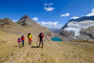 Gruppe wandert durch bräunliche Berglandschaft, im Hintergrund türkiser Bergsee und Gletscher