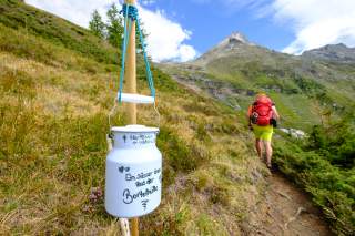 Milchkanne mit Bombons hängt in Berglandschaft, durch die eine Person wandert