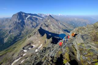 Vier Personen steigen am Seil Berg hinauf, weiter unten ein blauer Bergsee