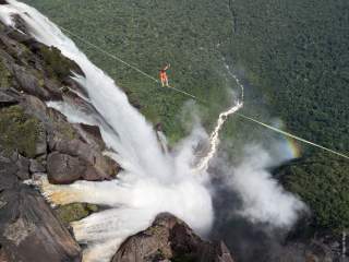 Eine Person balanciert auf einem Seil über einem hohen Wasserfall, darunter dichter Wassernebel und ein Regenbogen, im Hintergrund dichter Wald.