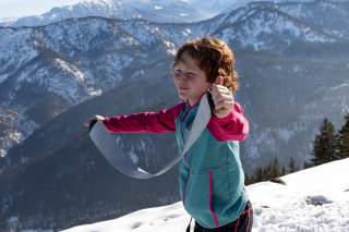 Kind steht vor winterlicher Bergkulisse und hat ein Skifell in der Hand.
