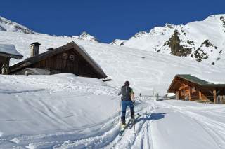 Skitourengeher zwischen zwei Hütten unter blauem Himmel.