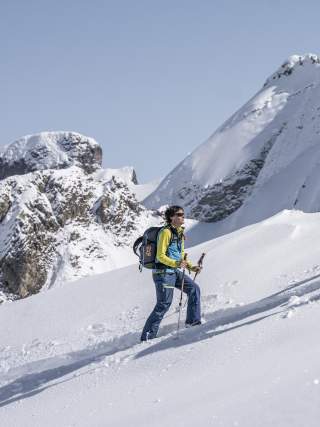 Ein Skitourengeher geht bei sonnigem Wetter mit Skitourenausrüstung einen verschneiten Hang hinauf, umgeben von schneebedeckten Berggipfeln.