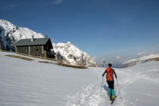 Person auf Tourenski läuft auf Berghütte in verschneiter Landschaft zu