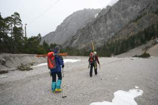 Zwei Menschen tragen Ski durch schotterigen Talboden